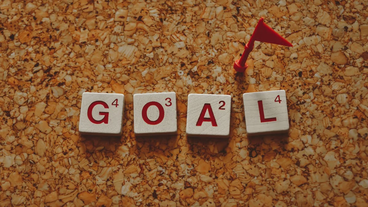 Scrabble tiles forming the word 'goal' with a small red flag on a cork board background.